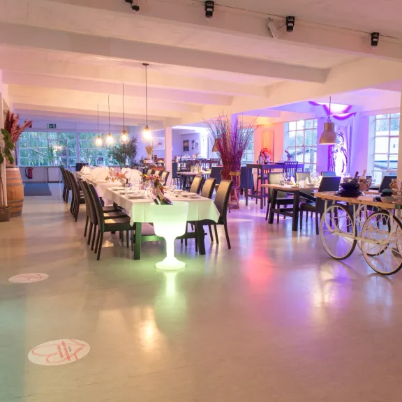 Modernes Restaurant mit bunt beleuchteten Tischen und Dekoration, helle Fenster im Hintergrund.