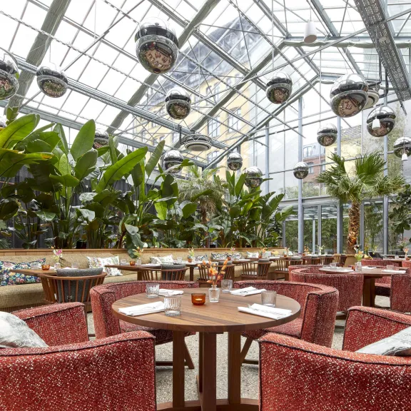 Restaurant mit runden Tischen, roten Sesseln und viel Grün unter Glasdach.