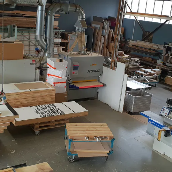 Holzwerkstatt mit Maschinen, Holzplatten und Werkzeugen, Tageslicht durch Fenster.