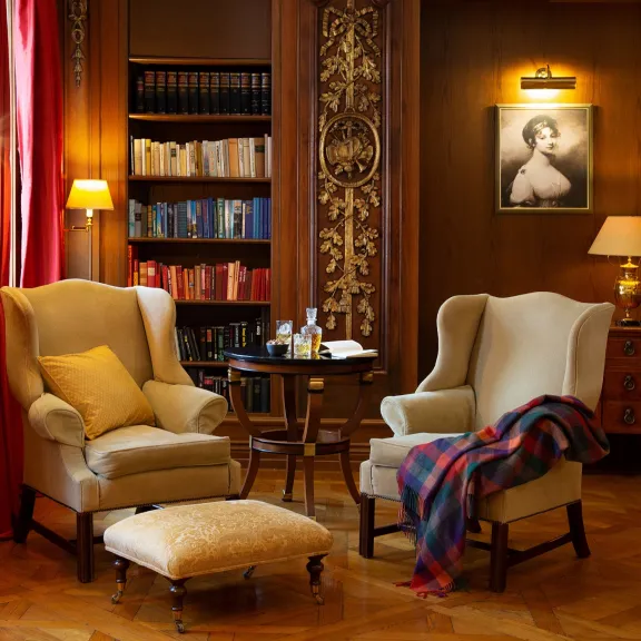 Gemütliche Bibliothek mit zwei Sesseln, rotem Vorhang, Bücherregal und warmem Licht.