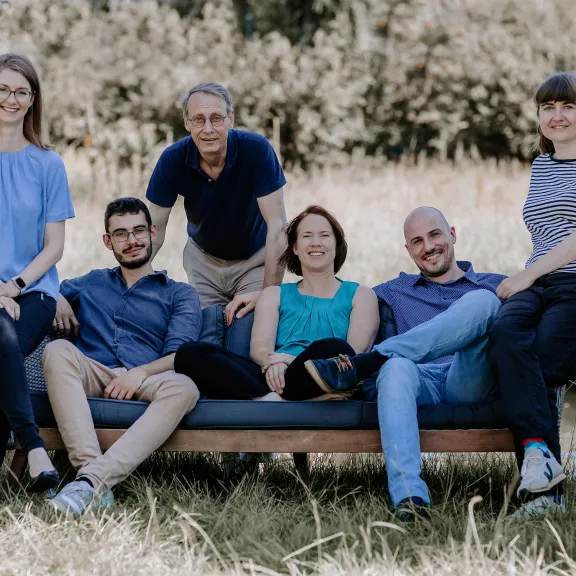 Sechs lächelnde Menschen sitzen und stehen auf einer Bank im Freien.