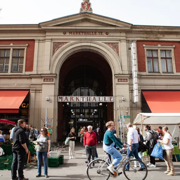 Menschen vor einem historischen Markthallengebäude mit roten Markisen und Ständen.