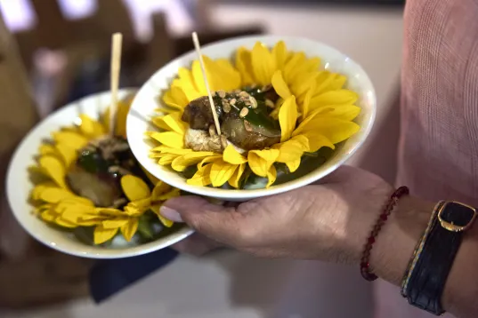 Hand holding two plates with food served on bright yellow artificial sunflowers.