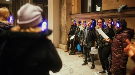 A group wearing illuminated headphones stands in an urban outdoor setting, performing a shared singing or team challenge during a Silent Disco Tour.