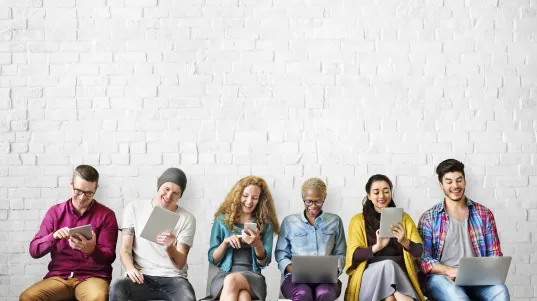 Six people sitting in a row, each using a digital device, against a white brick wall.