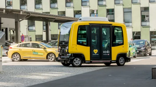 Yellow autonomous shuttle bus labeled “ioki” drives through a city street, with other vehicles and buildings in the background.