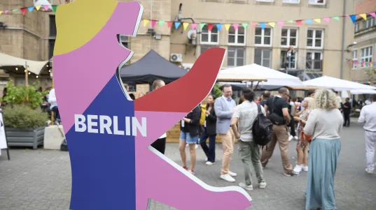 Colourful Berlin Bear sign in a courtyard with people and pennants at the BESTIVAL Festival. 