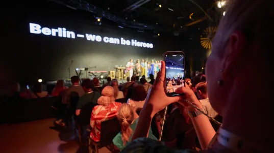 A stage with an LED screen displaying ‘Berlin – we can be Heroes’, an audience and a woman taking photos with her smartphone.