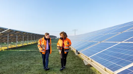 Male and female engineers working at solar power plant. Two technicians in reflective clothing walking between rows of photo…