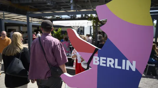Buntes Bären-Logo mit „Berlin“; Menschen gehen im sonnigen Außenbereich vorbei.