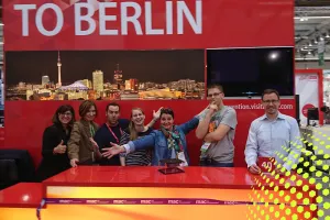 Sechs Menschen posieren fröhlich an einem roten Messestand mit Berlin-Stadtbild.