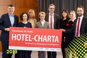 Acht Personen halten ein rotes Hotel-Charta-Banner, Jahreszahl 2019 unten links.