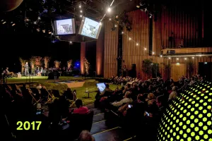 Publikum sitzt in einem dunklen Vortragssaal, Bühne mit Scheinwerfern beleuchtet, 2017.