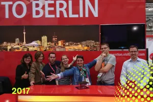 Sechs Personen lächeln an einem roten Messestand mit Berlin-Panorama im Hintergrund.
