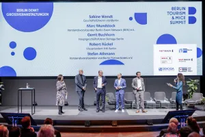 Six people stand on stage in front of a screen