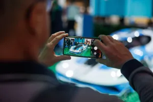 Person taking a photo of a blue sports car with a smartphone at an indoor event.