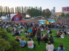 Crowd gathered on grass at an outdoor music festival with stage and large screens.
