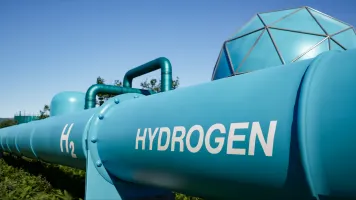 Large blue hydrogen pipeline labeled H₂ Hydrogen at an energy facility under a clear blue sky.