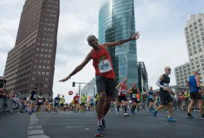 Runner in red balancing playfully during a city marathon, with tall buildings in the background.