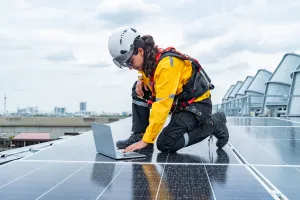 Technician checking solar panels on a rooftop using a laptop and safety gear.