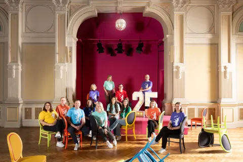 Team of Berlin Convention Office in colorful shirts pose on chairs in a grand hall with a large "25" sign.