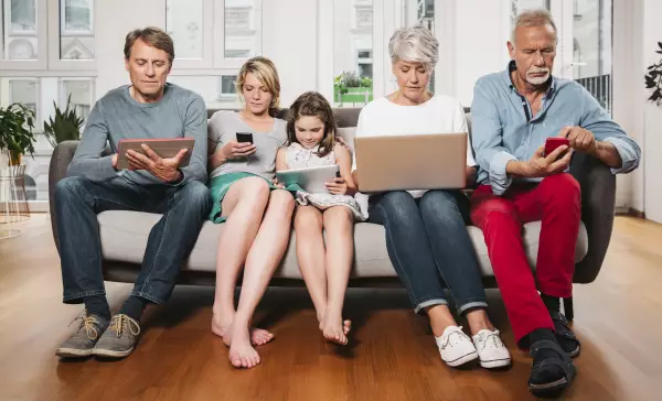 Group picture of three generations family sitting on one couch using different digital devices
