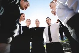 Sechs Menschen stehen lachend im Kreis, sonniger Tag, Teamstimmung.