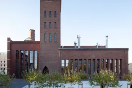 Backstein-Gebäude mit hohem Turm, davor Platz mit Bäumen und Sitzbänken.