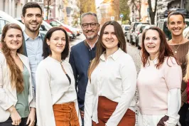 Neun lächelnde Menschen stehen auf einer sonnigen Straße in einer Stadt.