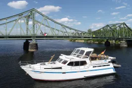 Weißes Motorboot auf Fluss vor grüner Stahlbrücke bei sonnigem Wetter.