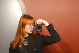 Frau mit rotem Haar macht ein Spiegelselfie vor roter Wand.
