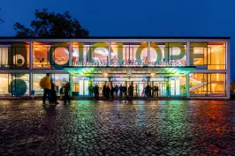 Modernes, bunt beleuchtetes Gebäude bei Nacht mit Menschen vor dem Eingang.