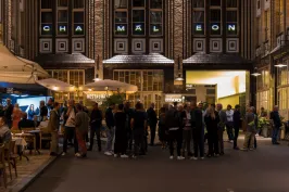 Menschenmenge vor einem beleuchteten Gebäude mit Schriftzug „Chamäleon“ am Abend.
