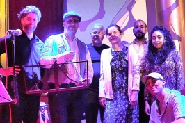 Gruppe von sieben Menschen auf einer Bühne, bunt beleuchtet, zwei halten Trommeln.