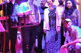 Gruppe von sieben Menschen auf einer Bühne, bunt beleuchtet, zwei halten Trommeln.