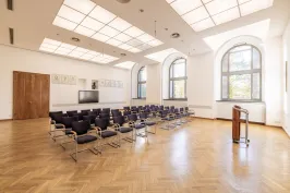 Heller, leerer Konferenzraum mit Stuhlreihen und Rednerpult, große Fenster und Holzboden.