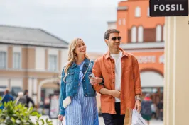 Paar spaziert lächelnd mit Einkaufstaschen durch eine Einkaufsstraße bei bewölktem Himmel.