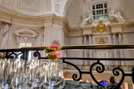 Mehrere leere Gläser und rosa Blumen vor prunkvoller, heller Saaldecke.