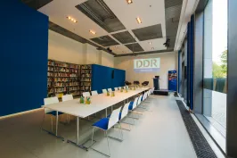 Moderner Konferenzraum mit langer Tafel, blau-weißen Stühlen und Bücherregalen.