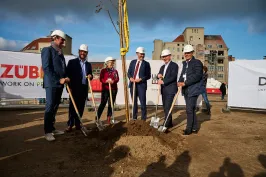 Sechs Personen mit Helmen pflanzen gemeinsam einen Baum auf einer Baustelle.