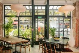 Modernes Café mit großen Fenstern, Holztischen, Pflanzen und hellen Lampen.