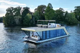 Modernes Hausboot mit Glasfront auf einem Fluss, umgeben von Bäumen.