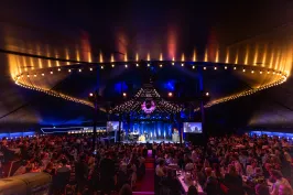 Der illuminierte, voll besuchte Saal im TIPI AM KANZLERAMT, Blick auf die Bühne davor Gäste an Tischen verteilt, nicht erkenntlich