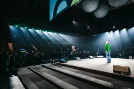 Person auf Bühne spricht vor großem, beleuchtetem Publikum in Arena.