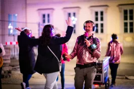 Moderator mit Mikrofon und Funkkopfhörern begleitet Teilnehmende einer Silent Disco Tour, die ebenfalls Kopfhörer tragen und im urbanen Außenraum interaktiv teilnehmen.