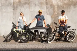 Drei Personen mit E-Bikes stehen vor einer Betonwand in der Sonne.