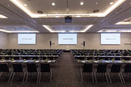 Großer, heller Konferenzraum mit vielen leeren Tischen und Stühlen, drei Leinwände vorne.