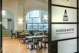 Modernes, helles Café mit Holztischen und Blumen, Eingang mit Logo „Hugo & Notte“.