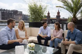 Fünf Menschen sitzen lachend auf einer Terrasse mit Blick auf die Oberbaumbrücke.