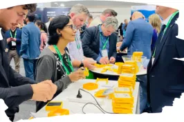 Menschen bei einer Messe, stehen an Tischen mit gelben Schachteln und Unterhaltungen.